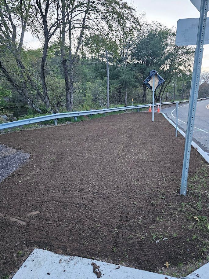 An empty patch of dark soil next to a road, bordered by a guardrail and sign.
