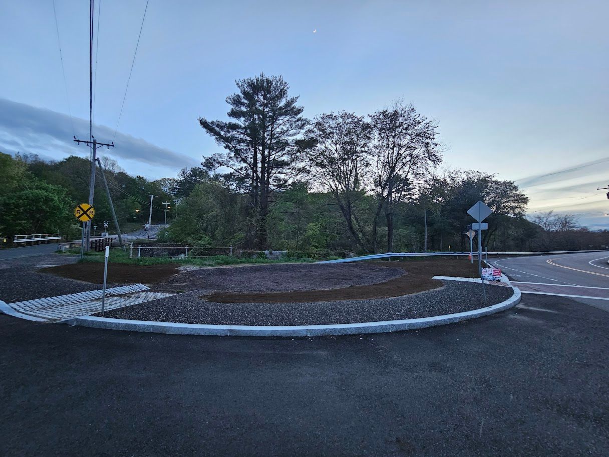 Newly constructed roundabout with dark asphalt, gravel border, and landscaping, under a cloudy sky.