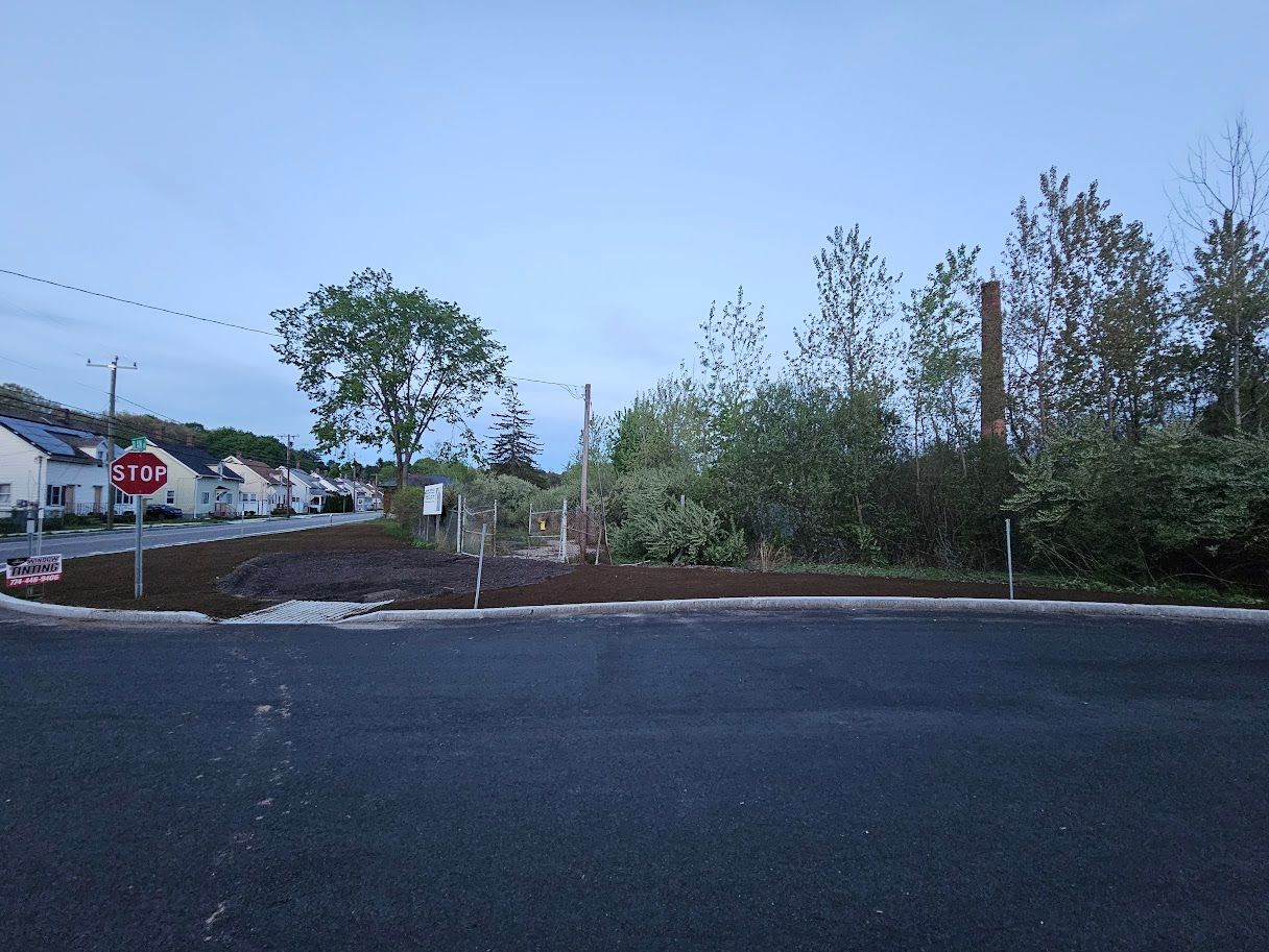A stop sign at a road corner, looking toward a fenced lot with trees and a smokestack in a residential area.