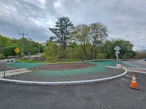 Roundabout with green and brown surface, trees in background, cloudy sky.