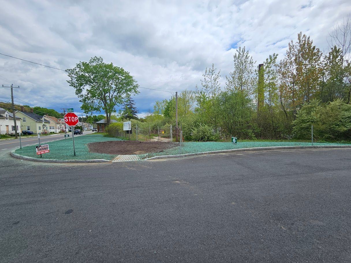 An empty lot with green-treated soil next to a road, stop sign visible, cloudy sky.