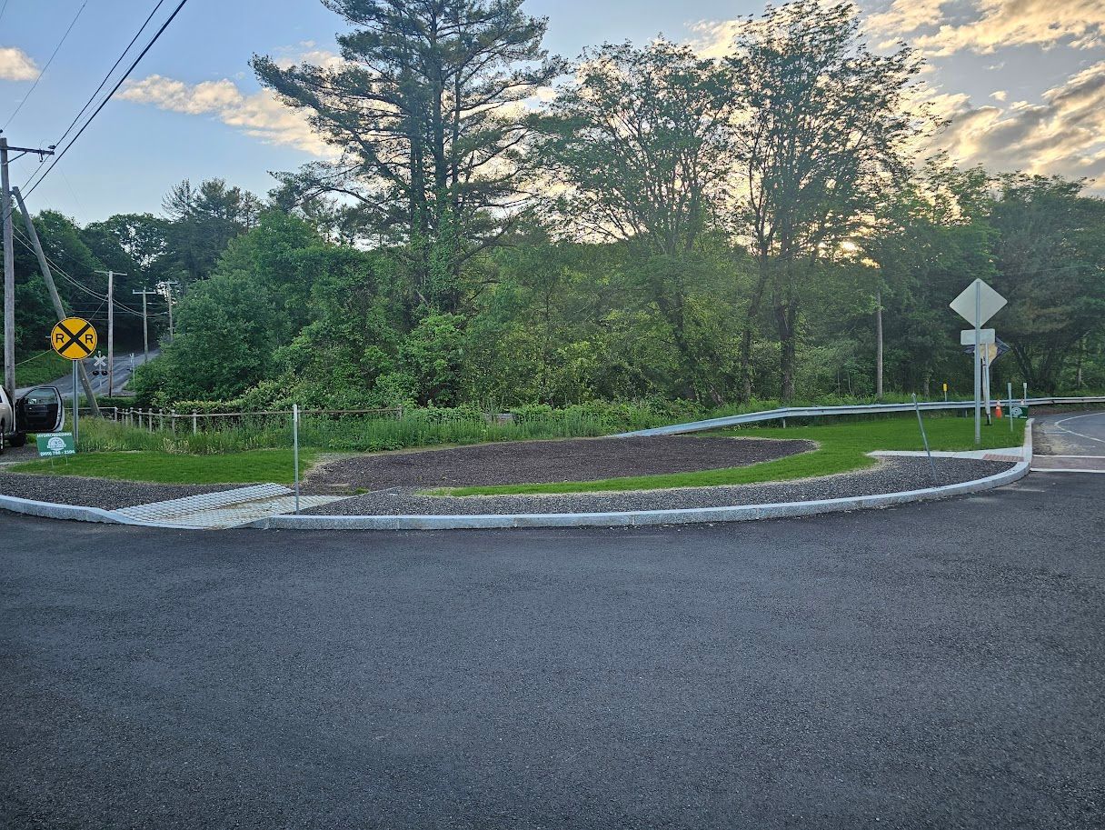 Newly paved road curves near a green space. Roadside guardrails and a railroad crossing sign are visible.