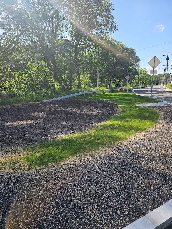 A gravel path leads to a grassy area with trees and road signs under a sunny sky.