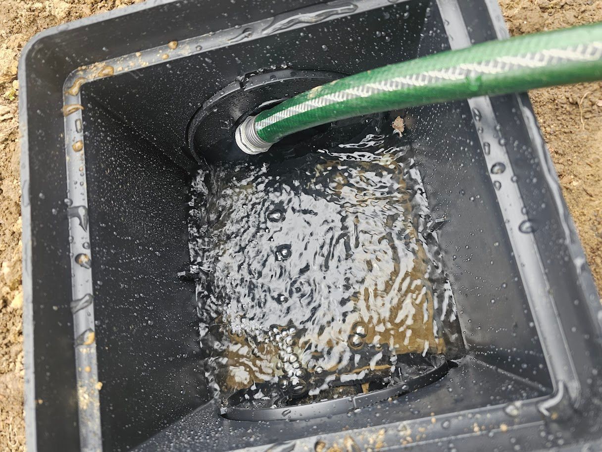 Hose spraying water into a black, square basin filled with murky water and debris.