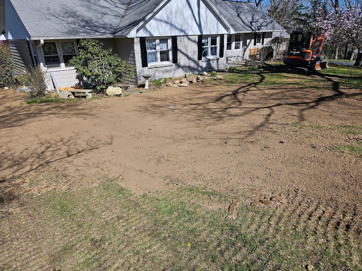 A house and dirt lot, tracks on grass. A small tractor is in the background. Sunny day.