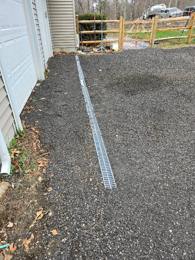 Gravel driveway with a long, metal drainage grate next to a garage; wooden fence in the background.