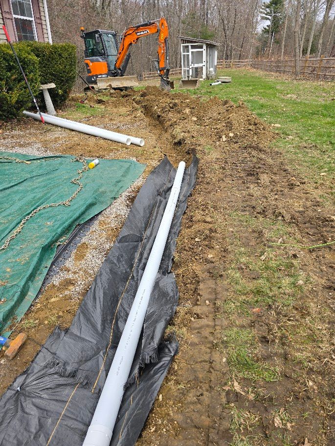 Trench dug in yard with black fabric and white pipe, orange excavator in background.