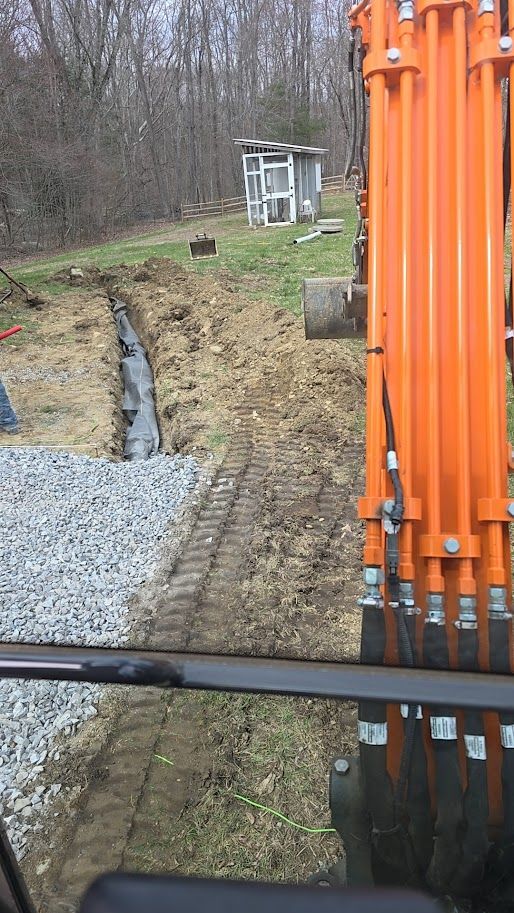 An excavator digging a trench near a wooded area. Orange arm, dirt, and gravel visible.