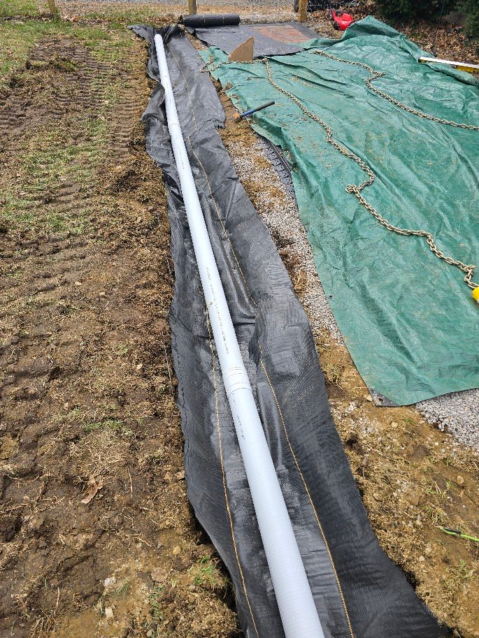 White perforated pipe laid in trench, wrapped with black fabric and gravel, outdoors.