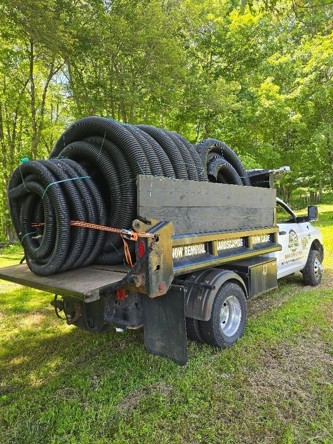 Truck bed loaded with black corrugated drain pipes, secured with straps. Outdoors, green grass and trees.
