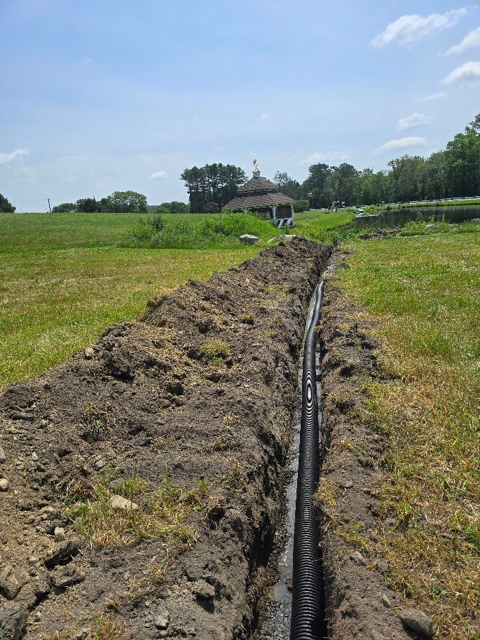 A narrow trench in a grassy field with black corrugated drainage pipe. A gazebo is in the background.