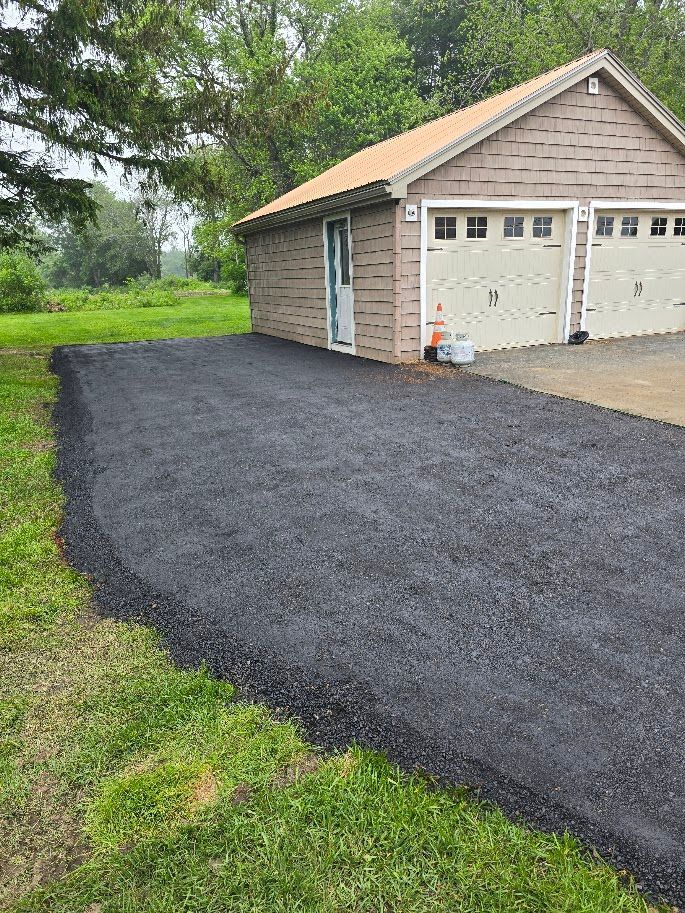 Newly paved asphalt driveway leading to a two-car garage with tan siding, next to green grass.