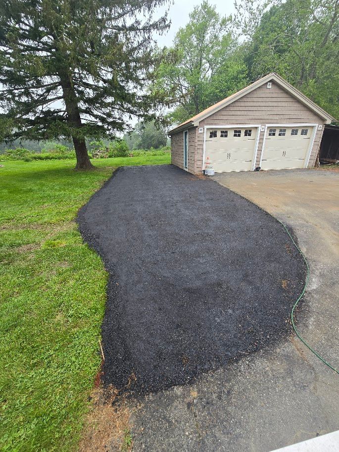 Newly paved black asphalt driveway leading to a two-car garage. Green grass and trees in the background.