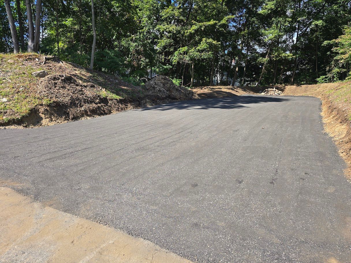 Asphalt driveway leading into a gravel parking area, surrounded by grass and trees.