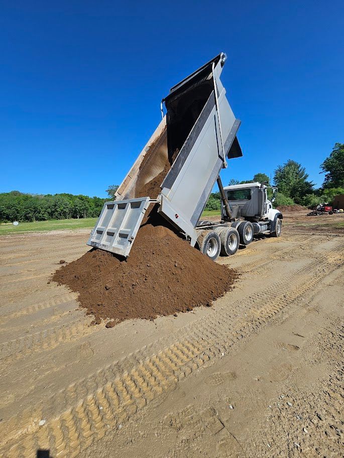 A white dump truck with its bed raised, unloading a large pile of brown soil onto a dirt ground under a clear blue sky
