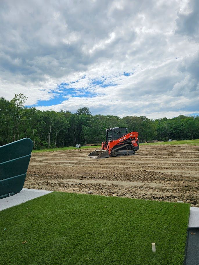 An orange skid steer loader sits on a dirt construction site in front of a tree line under a cloudy sky
