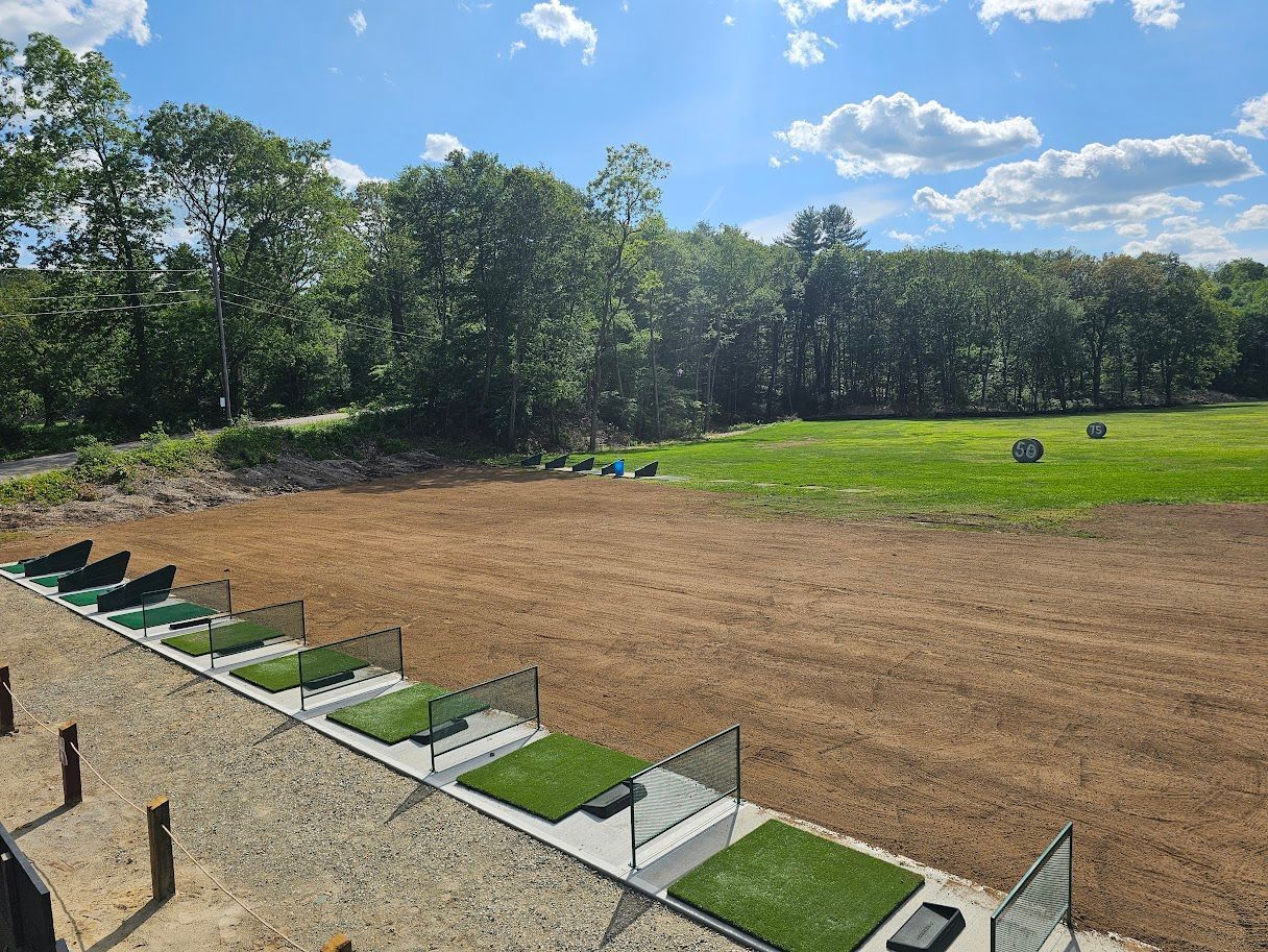 An outdoor driving range with multiple hitting bays featuring green mats, facing a large, dirt field and tree line