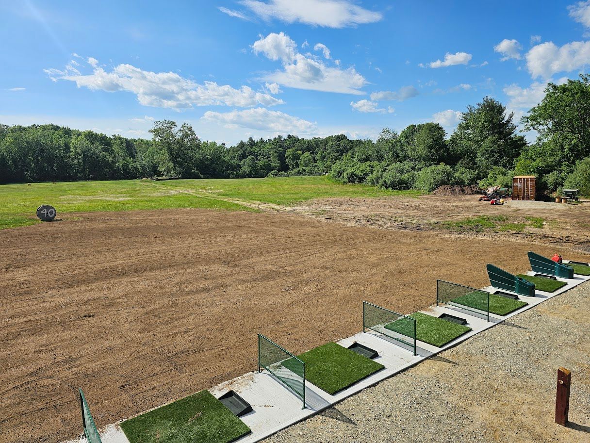 A driving range with multiple grass tee stations, a bare dirt landing area, and a lush green forest background