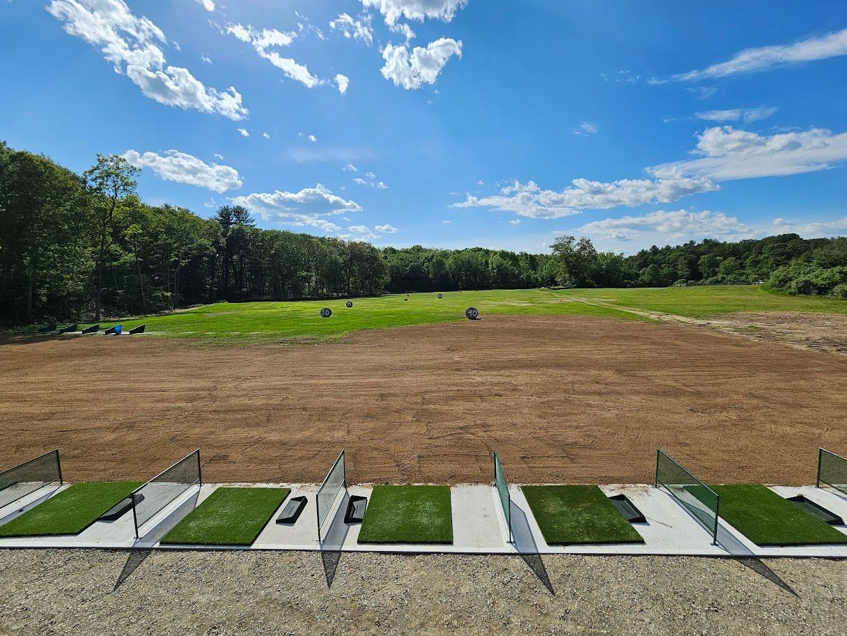 A driving range with multiple synthetic turf hitting bays separated by glass partitions, facing an open grassy field