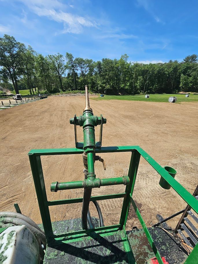 A green manual irrigation nozzle mounted on a metal frame, positioned over a tilled agricultural field under a blue sky