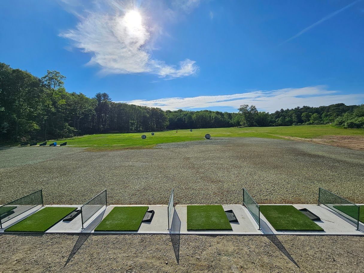 A golf driving range with four grass hitting mats separated by glass dividers, facing an open green field under blue sky