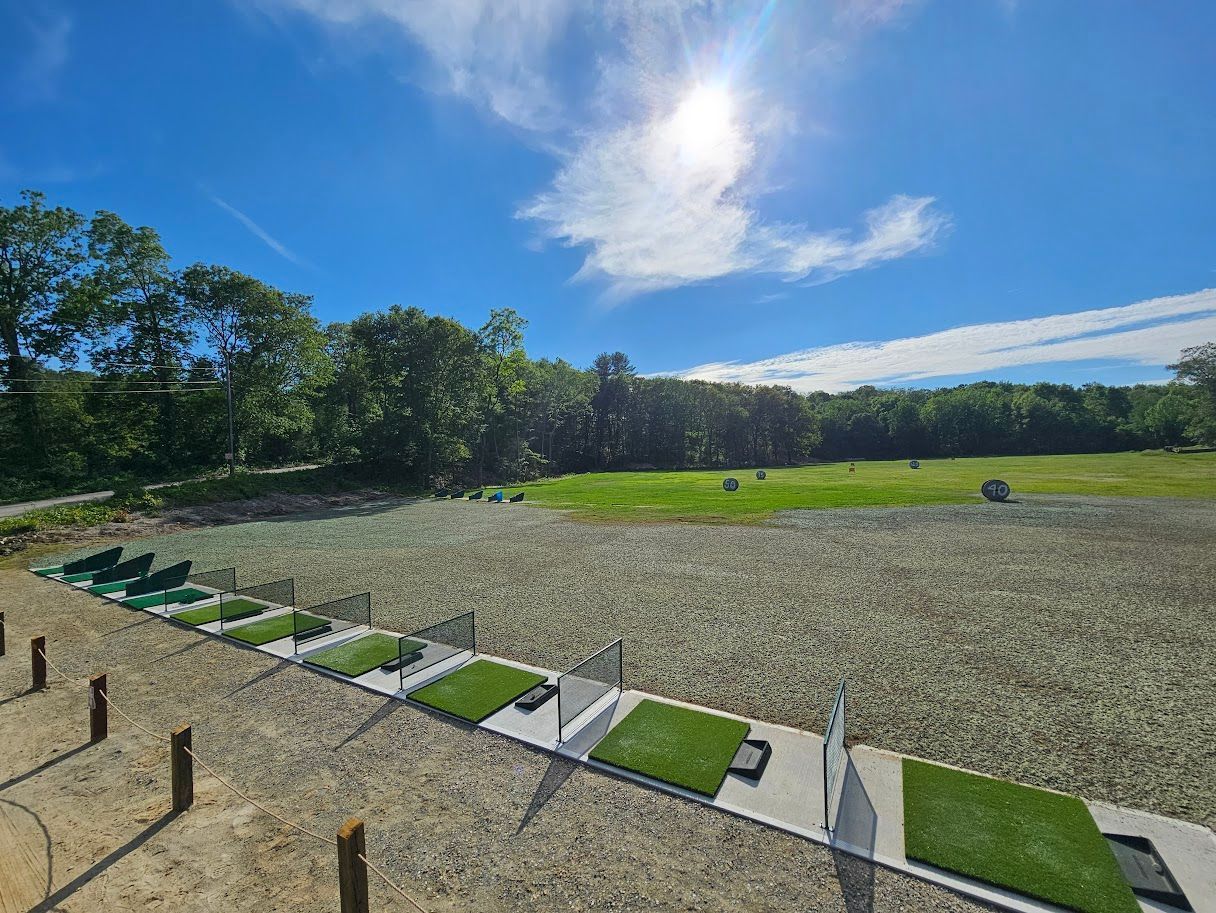 An outdoor driving range with multiple hitting bays featuring green turf mats, lined up along a gravel area under a sun