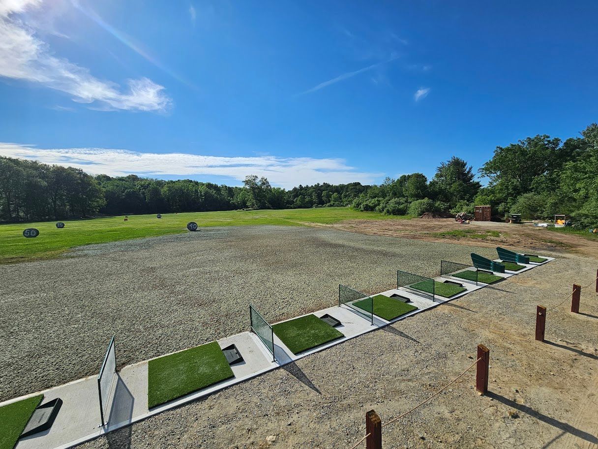 A driving range featuring a row of matted hitting bays with dividers, facing a large, open field under a blue sky