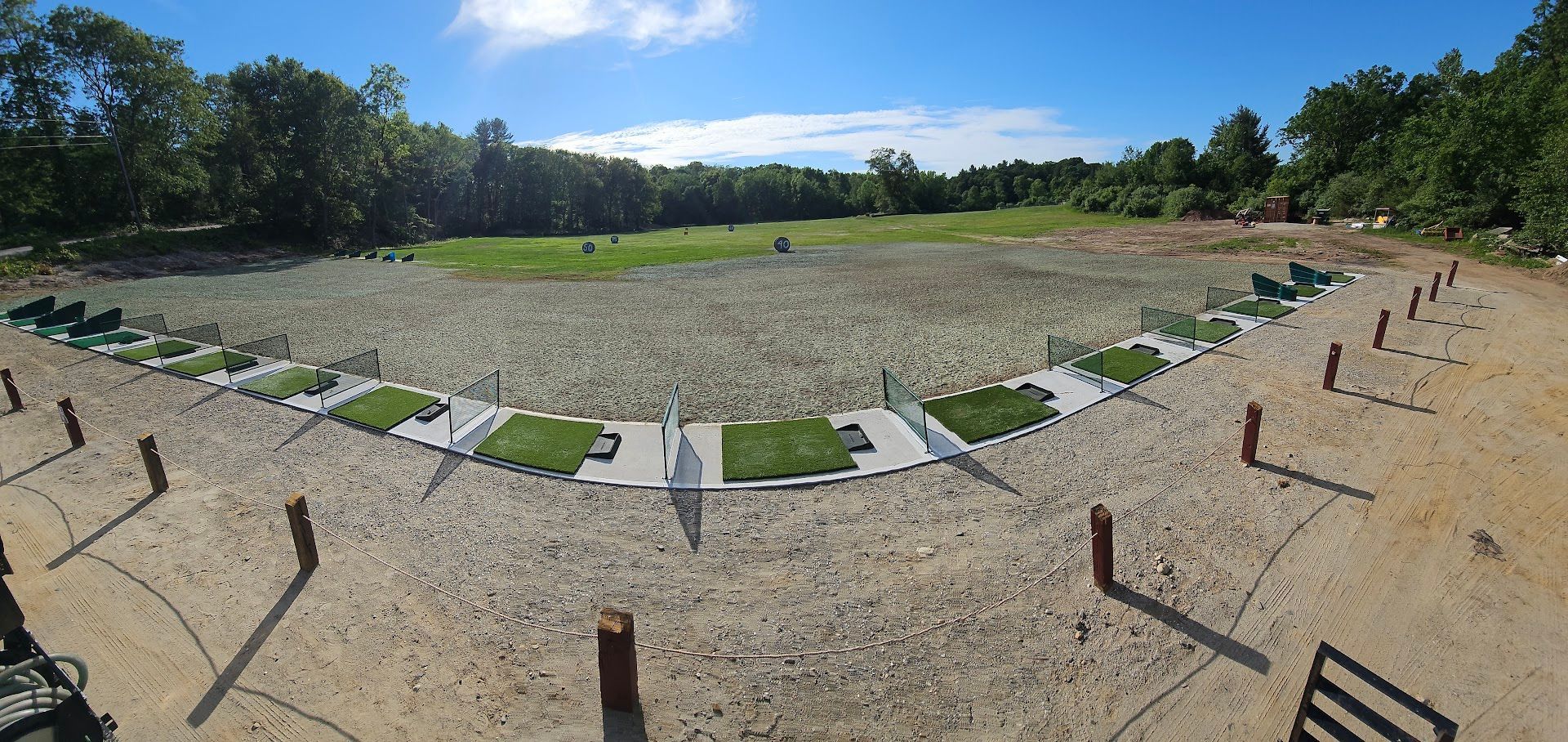A wide-angle view of a golf driving range featuring multiple grass hitting bays and a large, open field under a blue sky