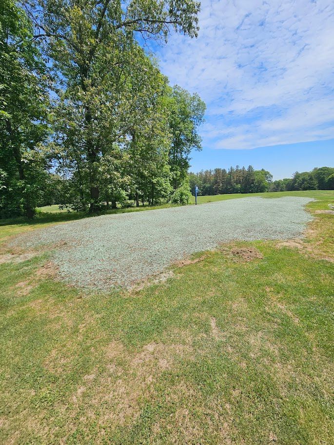 A patch of gravel sits in a grassy field bordered by trees under a blue, partly cloudy sky