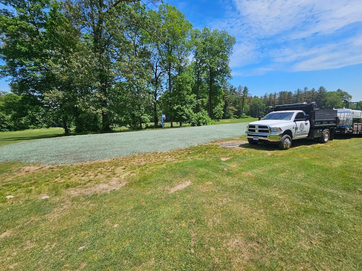 A white pickup truck with a trailer is parked on a grassy field next to a large area covered with light gray gravel