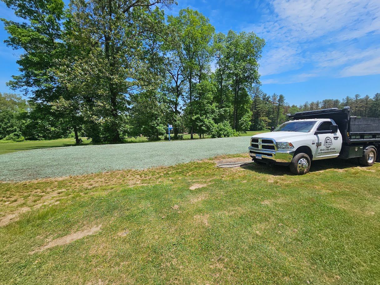 A white dump truck parked on a grassy field next to a large area covered in spread gravel near a line of trees