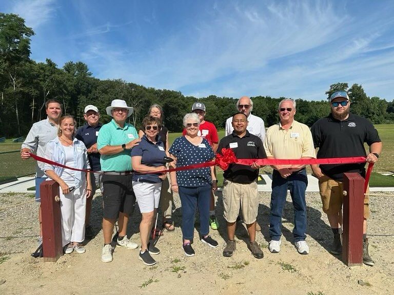 A group of people standing outdoors at a ribbon-cutting ceremony, holding a red ribbon across a gravel path