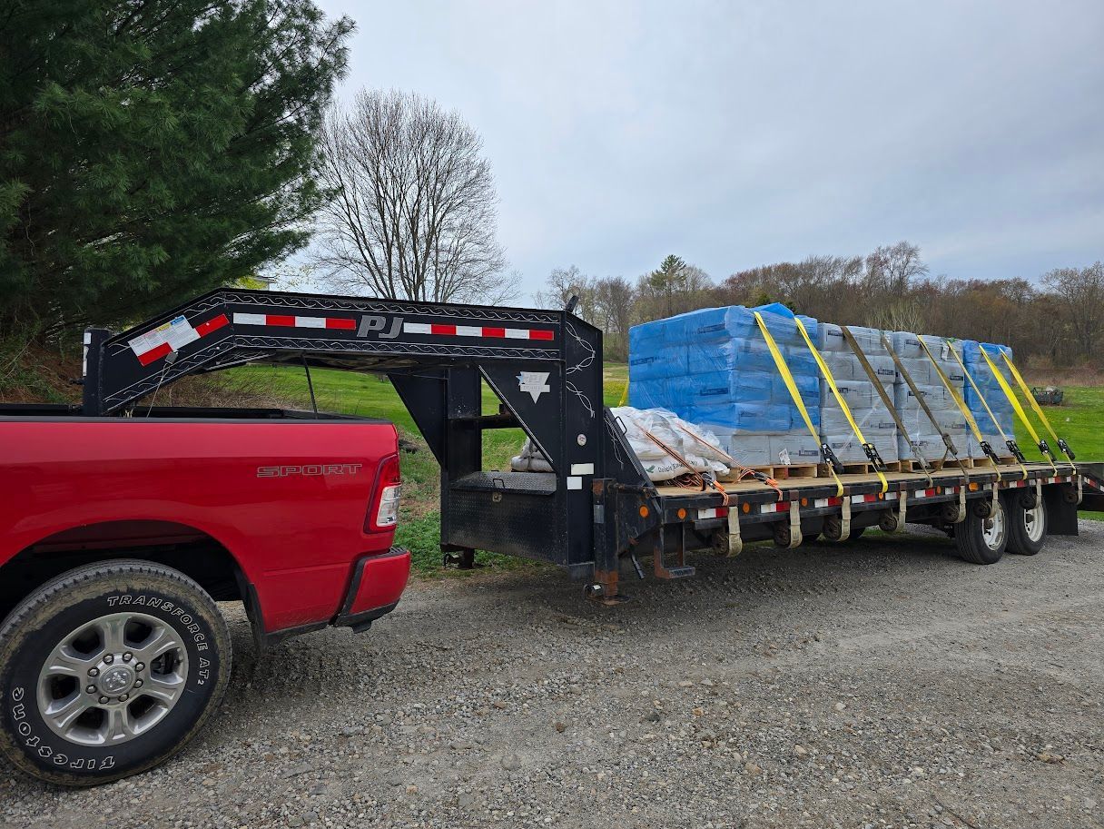 Red pickup truck towing a flatbed trailer loaded with wrapped construction materials, secured with straps.