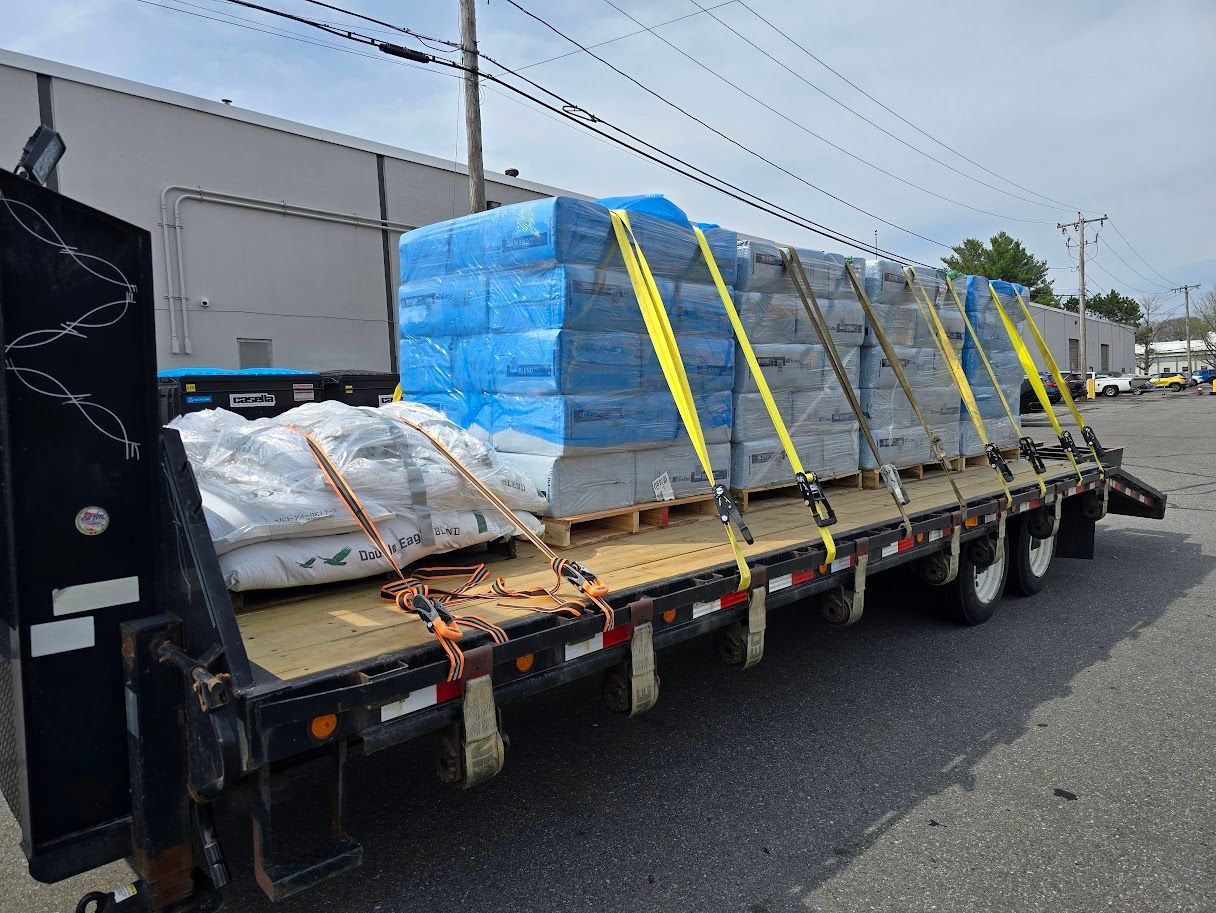 Flatbed truck loaded with wrapped pallets of building materials, secured with straps. Sunny day.