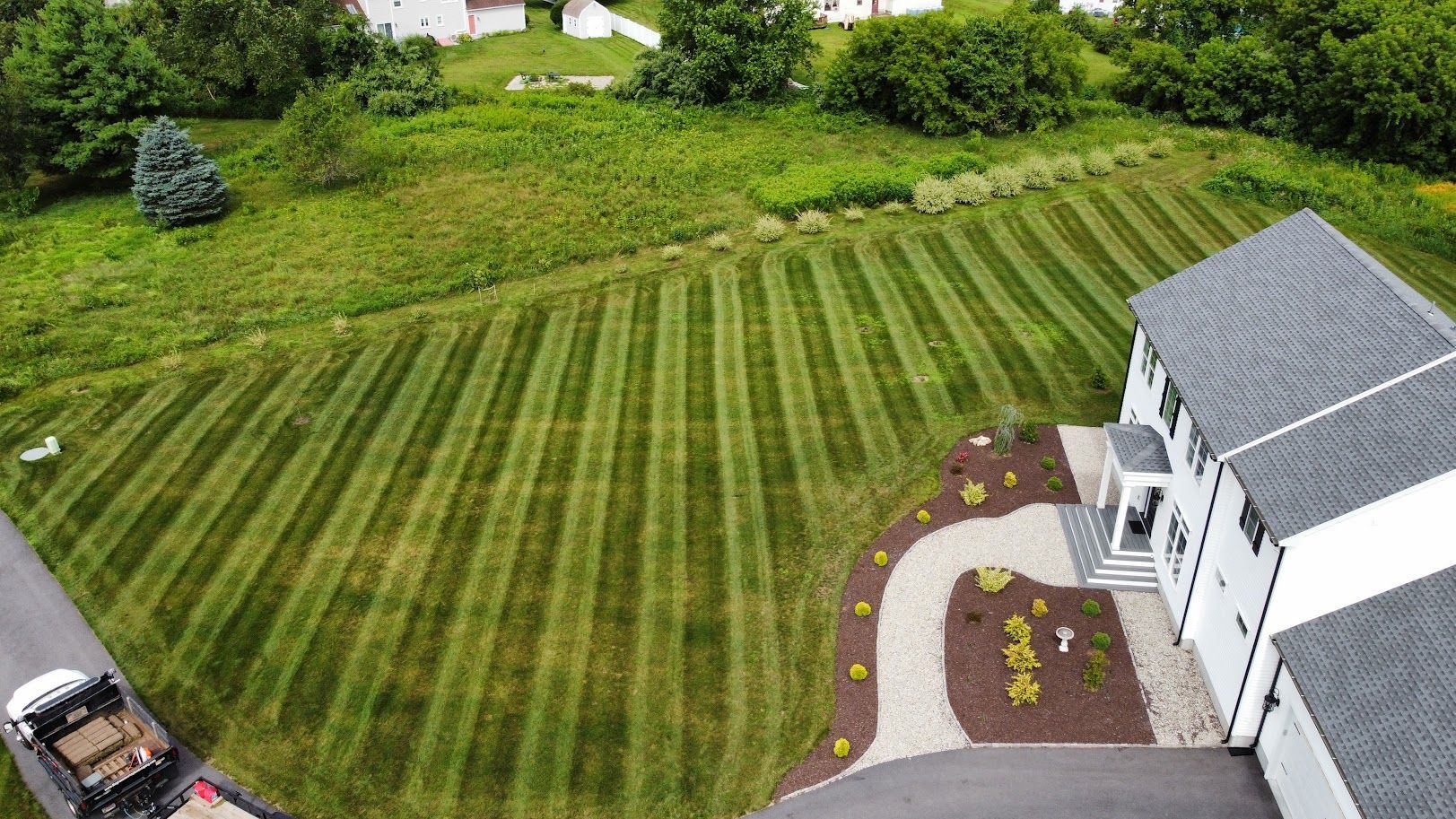 Drone view of a house with freshly mowed lawn, dark gray roof, and stone walkway. A landscaping truck is parked nearby.