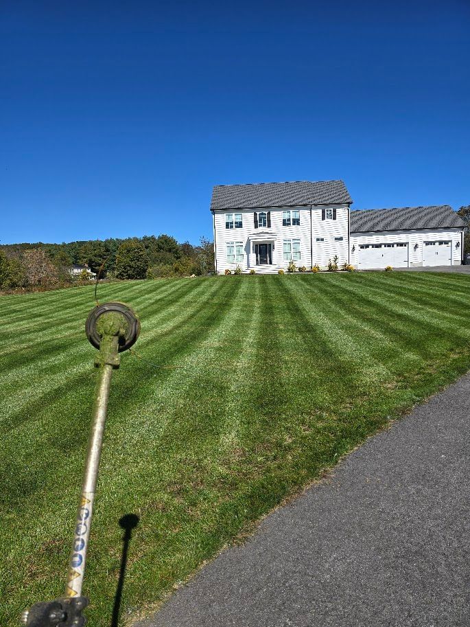 String trimmer on a striped lawn, large white house in background under a blue sky.