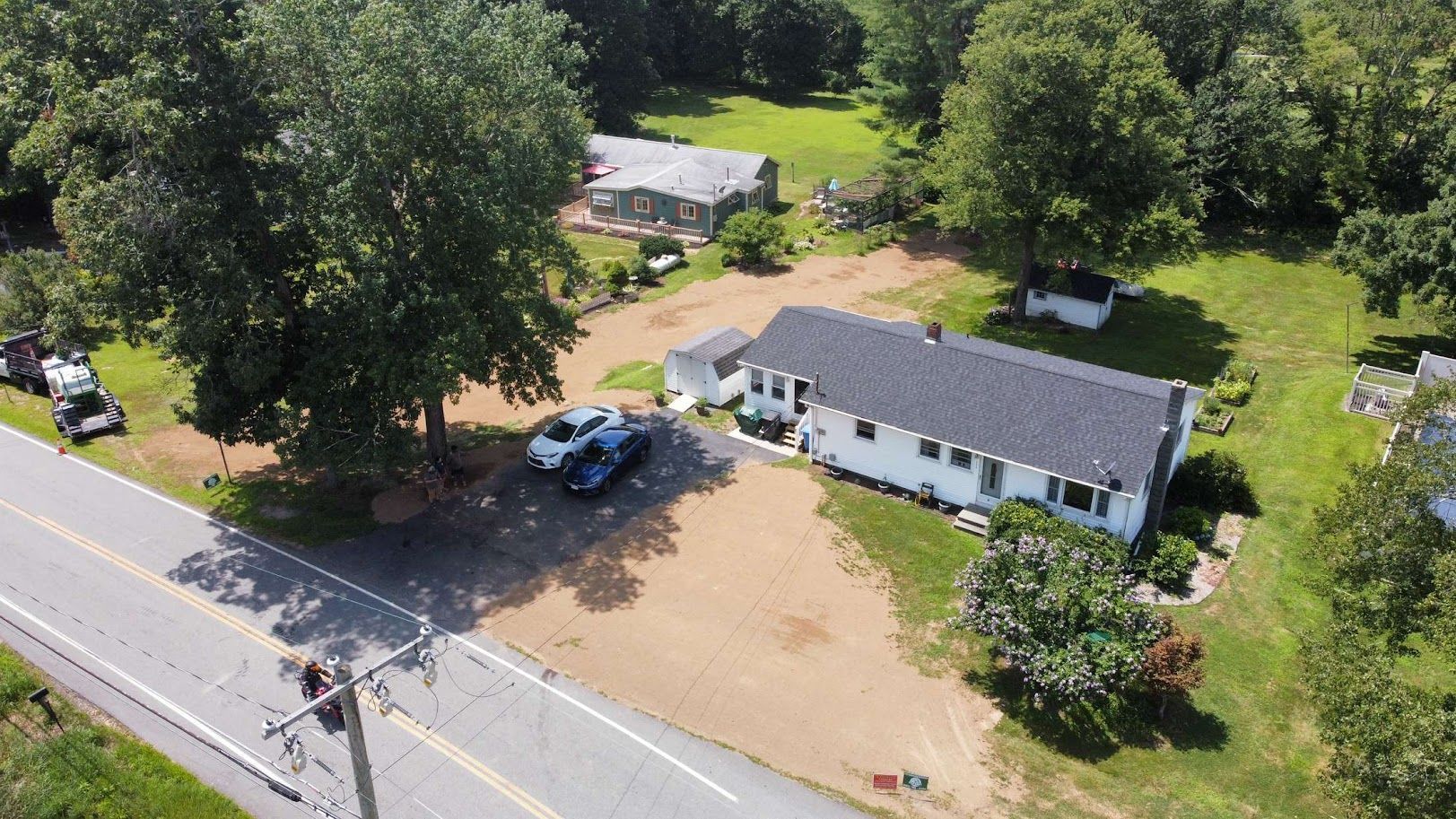 Aerial view of a white ranch-style house with a dark roof, adjacent to a road and large trees in a rural setting