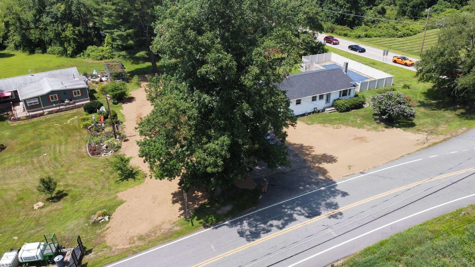 Aerial view of two houses surrounded by green trees and fields, situated next to an asphalt road with cars