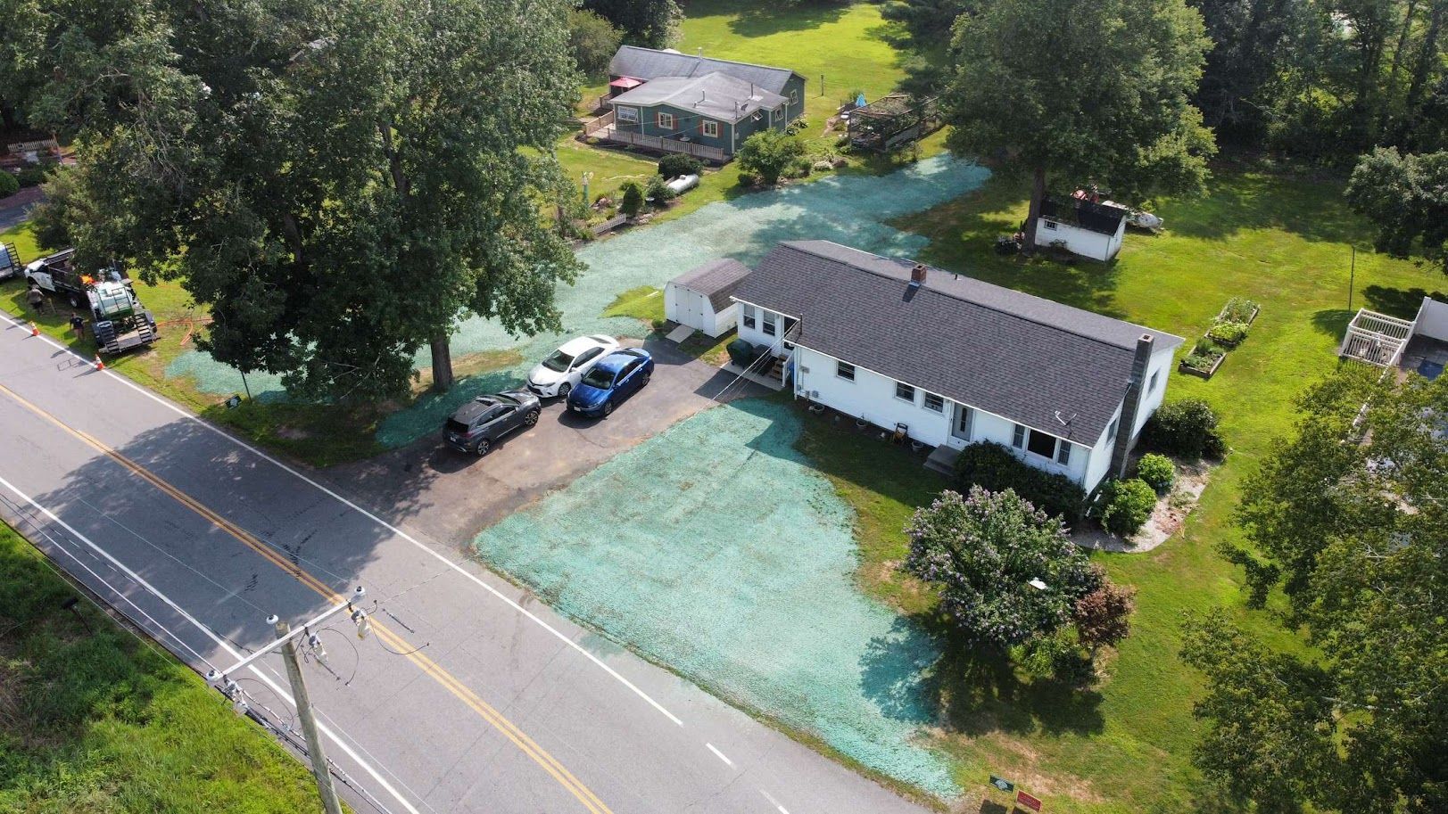 An aerial view of a white, single-story house with a dark roof, adjacent to a paved road with cars parked in the driveway