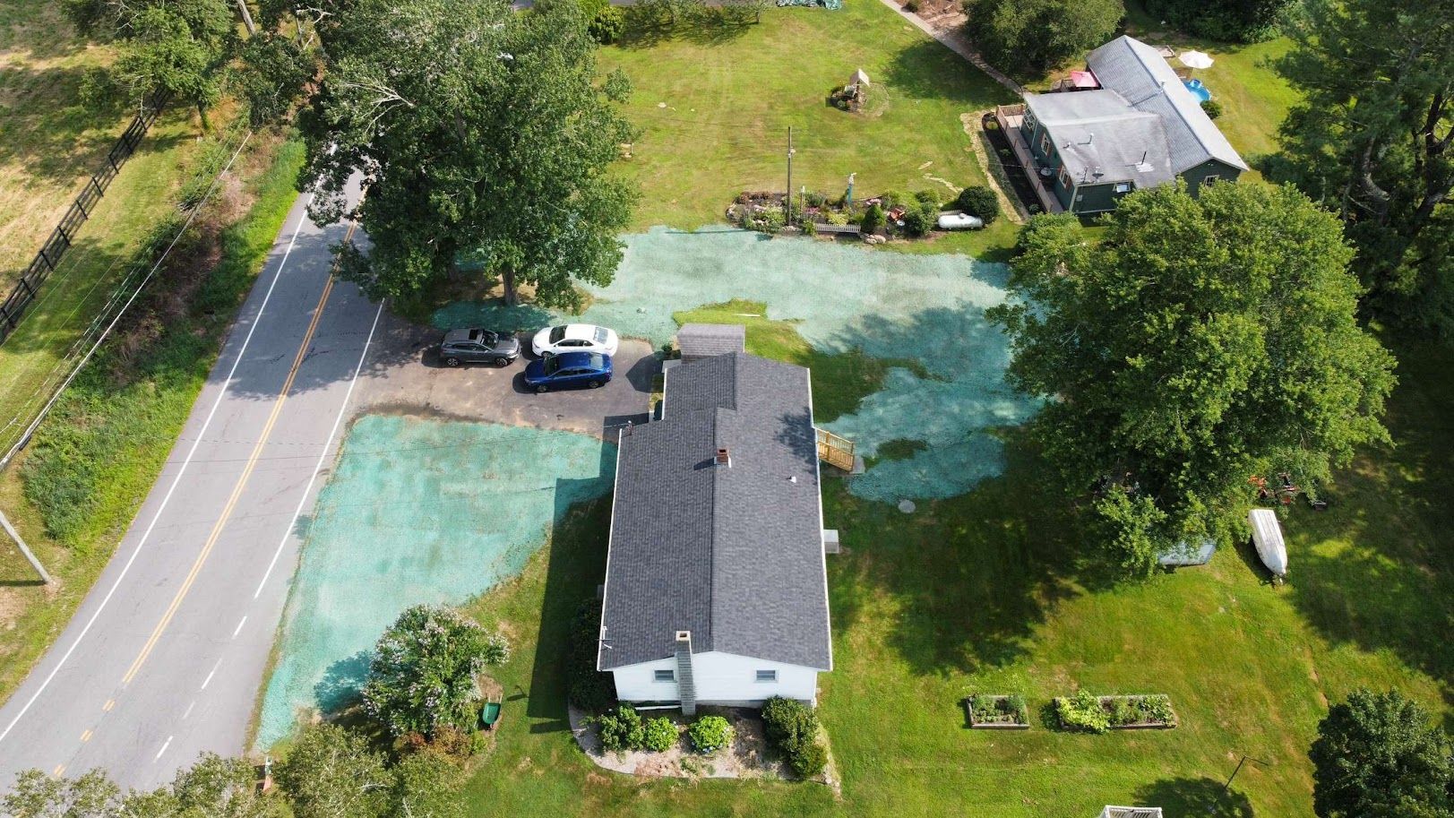 Aerial view of a house with a dark roof and a neighboring detached garage, surrounded by grass and trees near a road
