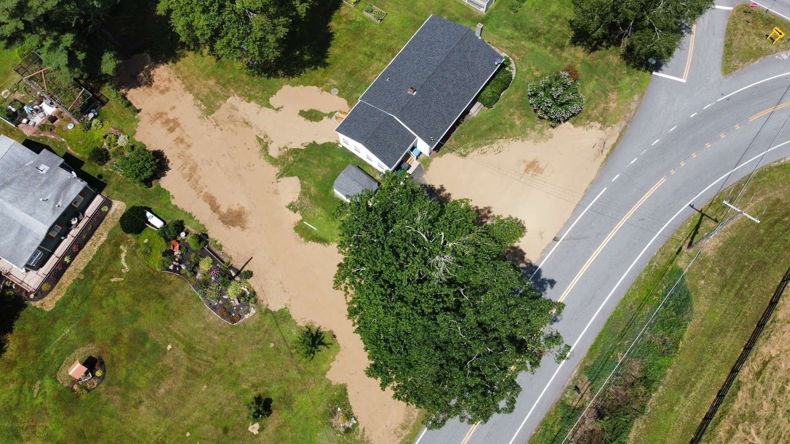 An aerial view shows muddy floodwater covering a yard and driveway next to a house and a road