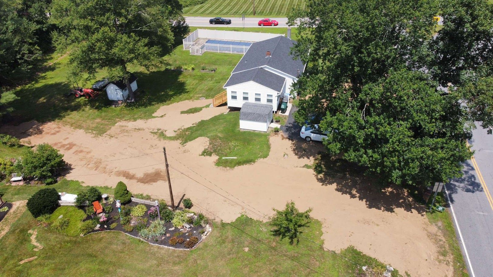 Aerial view of a white single-story house with a black roof, large trees, and a dirt yard next to a paved road