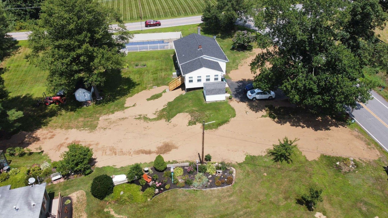 An aerial view of a house surrounded by a large area of bare, brown dirt and scattered trees, with a road nearby