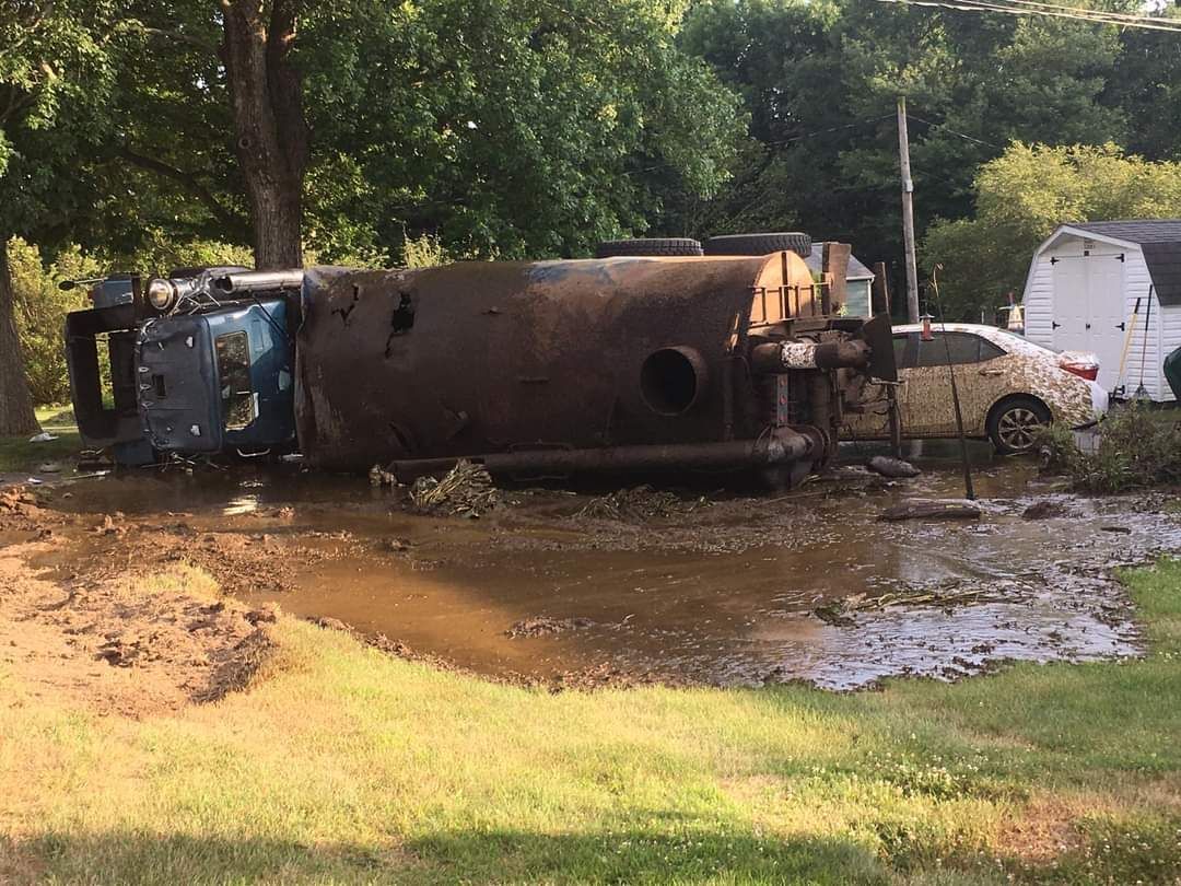A large, dark tanker truck lies on its side in a muddy, grassy area near a white car and a shed