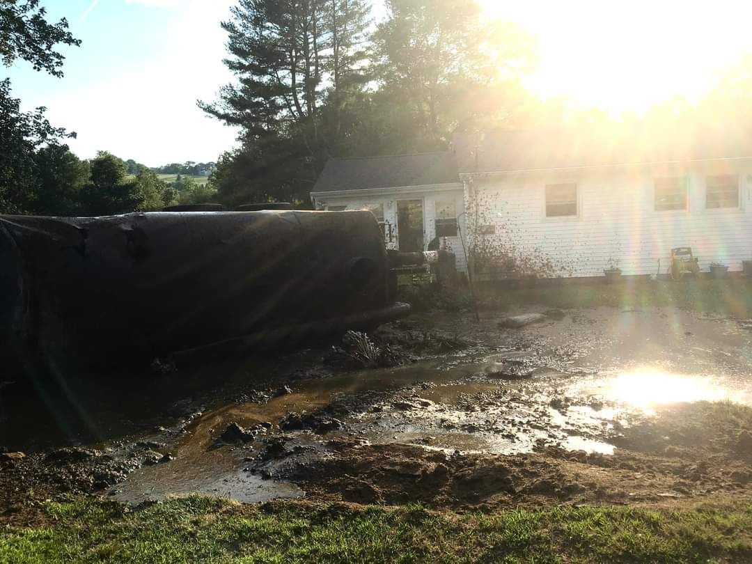 A flooded yard with muddy water near a white house at sunset, with a large, dark structure partially submerged