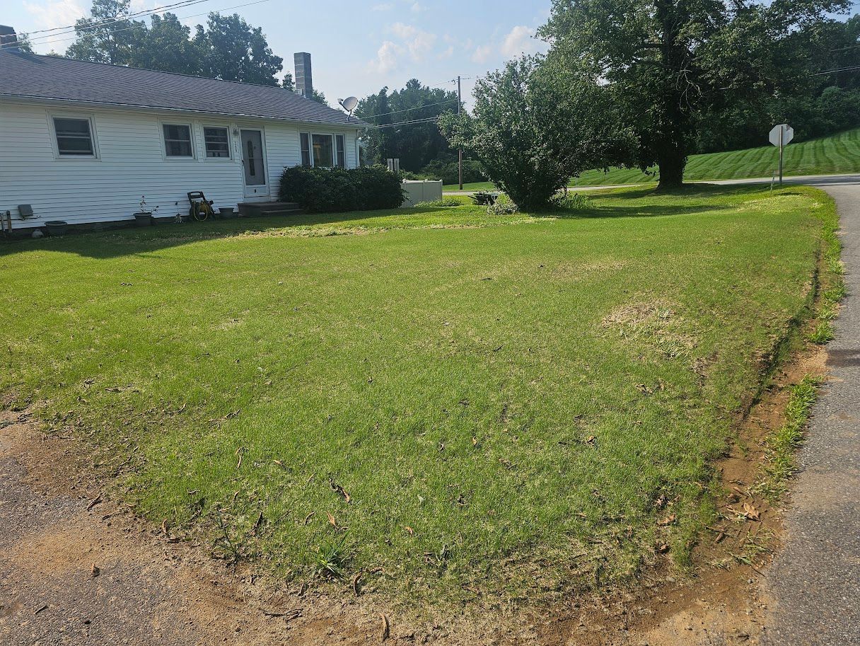 A white house with a metal roof sits behind a large, grassy yard adjacent to a paved road on a sunny day