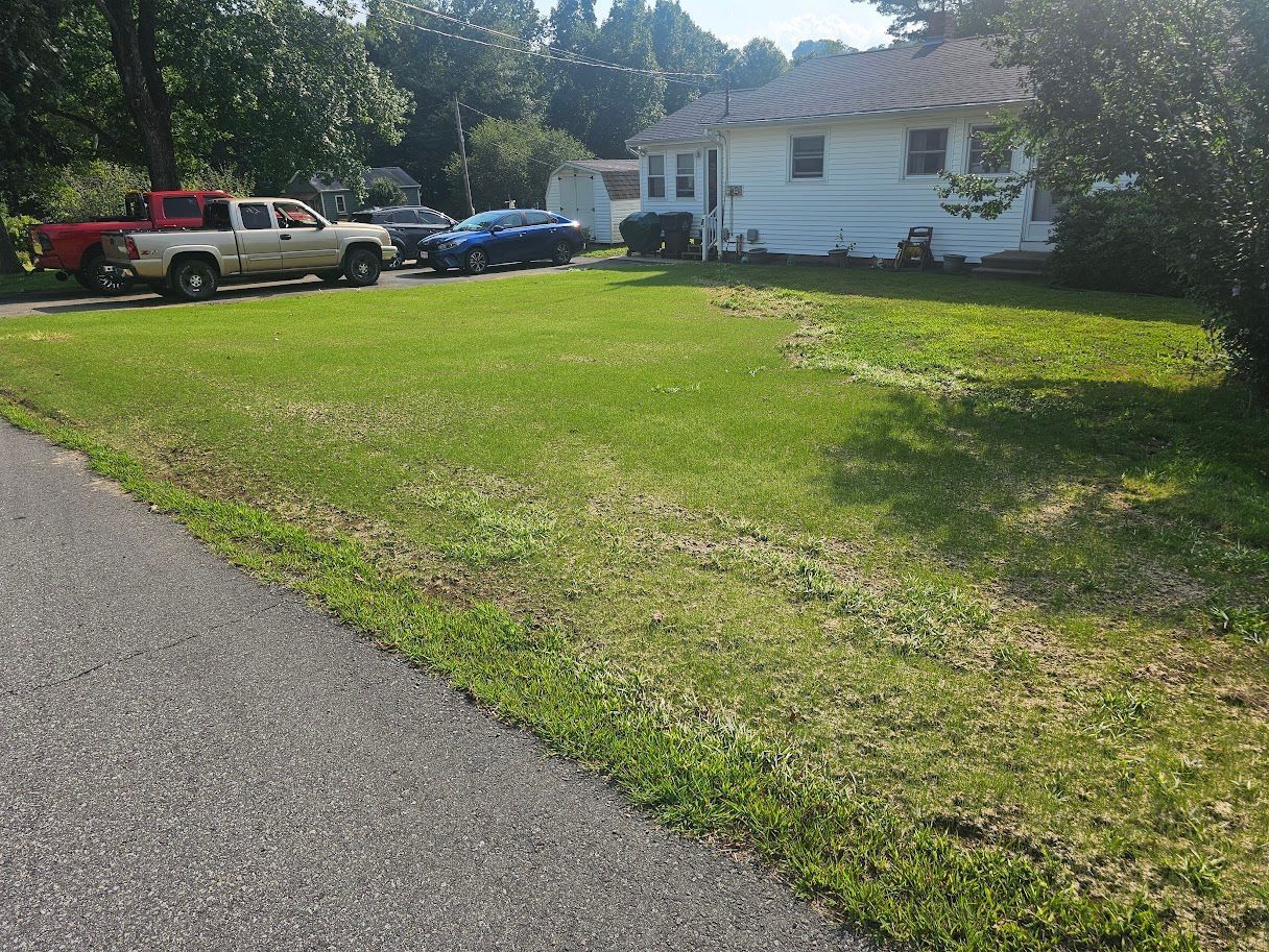 A white house sits behind a large green lawn, with a paved road in the foreground and several vehicles parked to the side