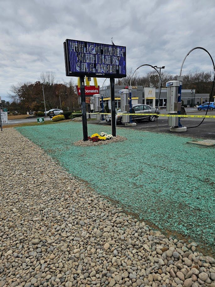 A car wash entrance with a dark digital sign, yellow caution tape, and a small memorial of flowers at the base of the sign