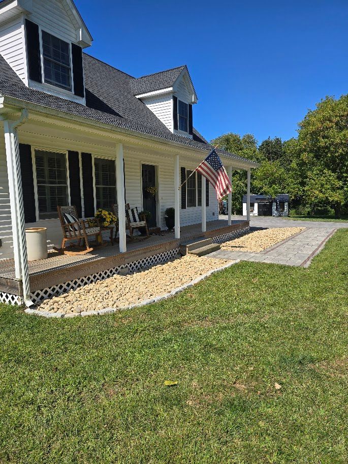 White house with porch, American flag, rock landscaping, green lawn, clear blue sky.