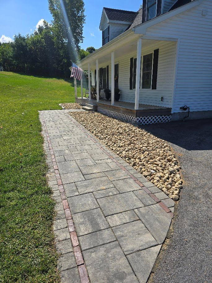 Paved walkway leading to a white house with a porch, bordered by rocks and grass.